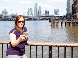 Manuela-Willbold-looking-at-her-mobile-phone-on-pier-with-London-skyline-in-background